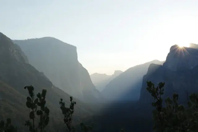 Morning sun breaking over a cliff on the right of Yosemite Valley, El Capitan in shadow on the left, haze filling the valley.