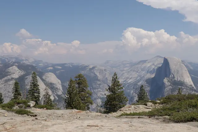 Half Dome and the high country under summer clouds, seen across granite slabs and scattered pines.