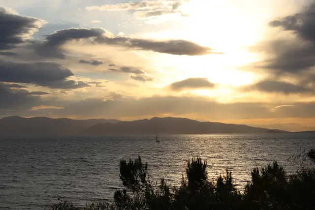 Sun breaking through heavy cloud above a calm sea, a sailboat and distant mountains, shrubs in silhouette in front.