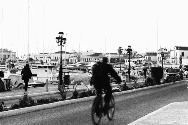 Black and white: a cyclist riding along Aegina's harbor road past lampposts, moored sailboats and white houses behind.
