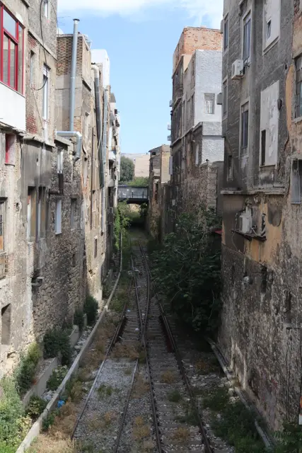 A narrow overgrown alley between weathered stone buildings, disused rail tracks running down its middle.