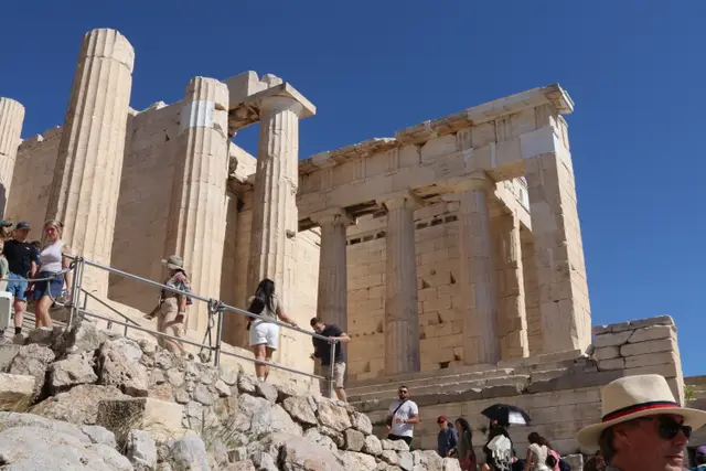 Visitors climbing the rocky path to the columned Propylaea gateway under a deep blue sky.