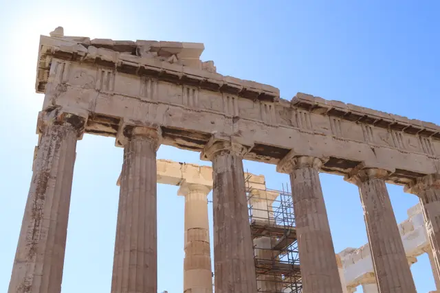 Looking up at the Parthenon's columns and entablature, scaffolding inside, the sun just out of frame.