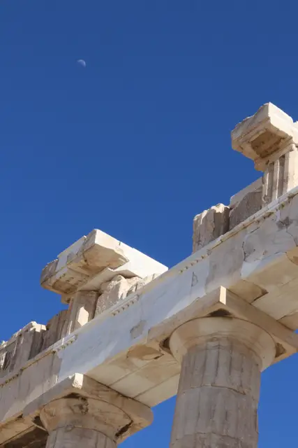 A corner of the Parthenon's marble cornice against a deep blue sky, a small half moon above.