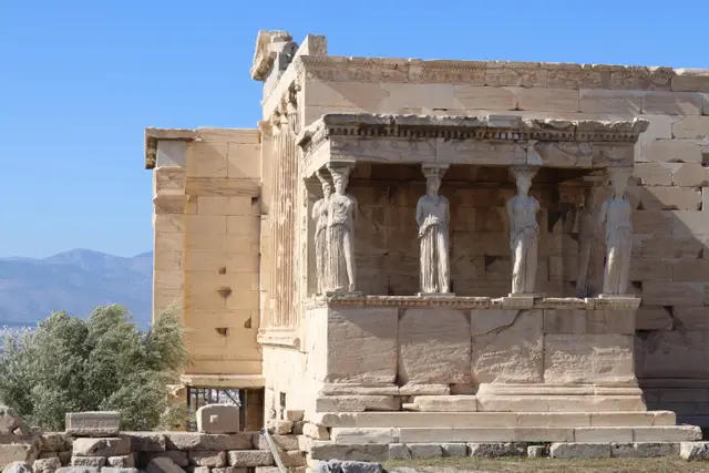 The Porch of the Caryatids on the Erechtheion, six stone maidens holding up the roof, an olive tree and hills behind.