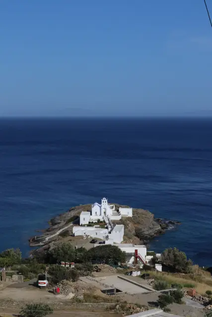 A small white monastery on a rocky islet below, a deep blue sea stretching to the horizon.