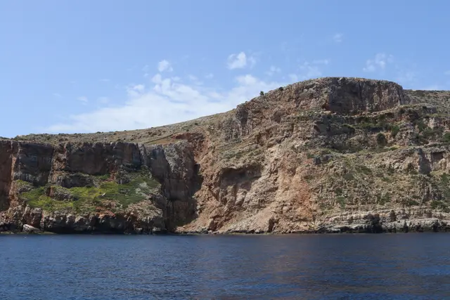 Red and ochre sea cliffs rising from dark blue water under a pale sky.