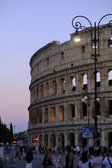 The Colosseum's outer wall at dusk under a lilac sky, a street lamp in front and crowds below.