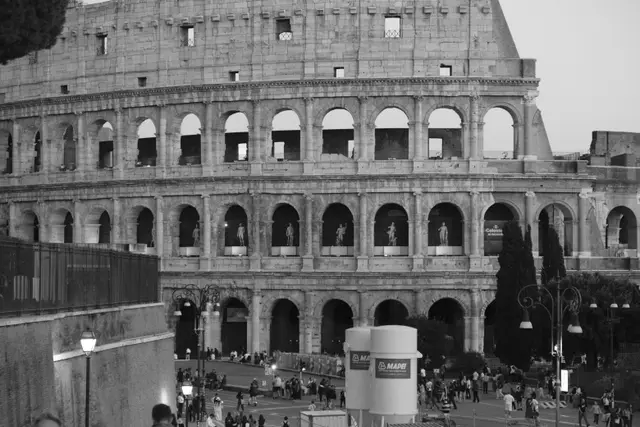 The Colosseum's arches in black and white, crowds and street lamps at its base.