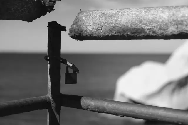 A padlock on a rusted seawall railing, the sea and a pale rock behind.