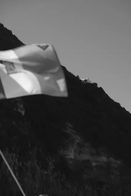 A flag blowing on the boat in front of a steep wooded cliff.