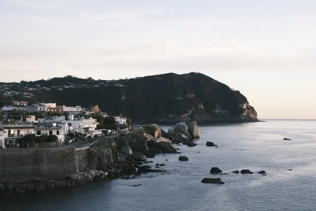 Forio's seafront in soft evening light, rocks in the water below a dark headland.