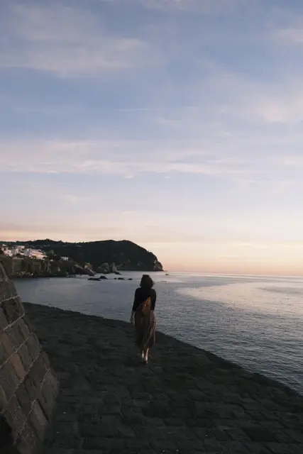 A figure walking along the curved stone seawall at dusk, a headland behind.