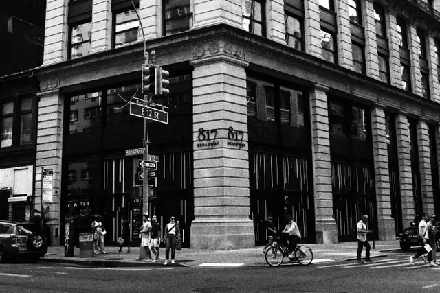 Black and white: the corner of a stone office building at East 12th Street, people crossing and a cyclist passing.