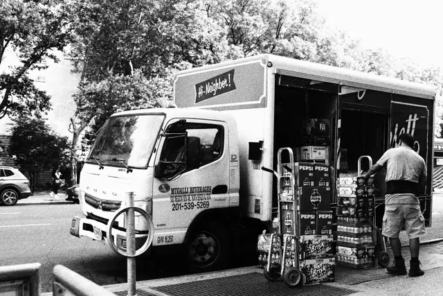 Black and white: a delivery truck parked on a tree-lined street, a man unloading cases of drinks.