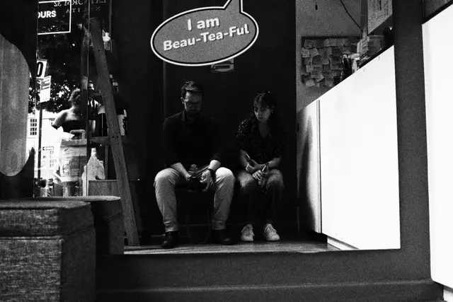 Black and white: two people sitting in a shop doorway under a speech-bubble sign reading I am Beau-Tea-Ful.