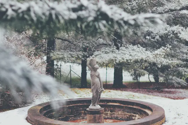 A small statue on a stone fountain, snow on the ground and pine branches around it.