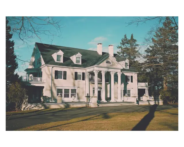 A white colonial house with dark shutters and a columned porch, tall evergreens beside it under a blue sky.
