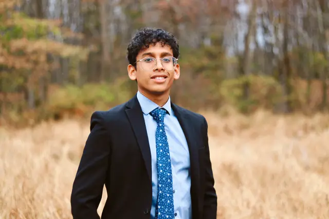 A young man in glasses, a dark suit and a blue patterned tie standing in tall golden grass, autumn trees behind.