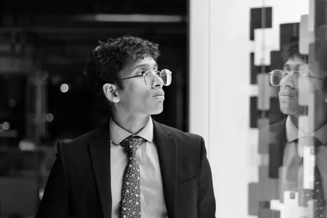 Black and white: a young man with curly hair, glasses and a patterned tie looking to the side, his reflection in a mirrored panel.