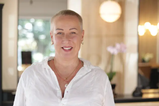 A smiling woman with short blond hair in a white shirt, a softly lit salon behind her.