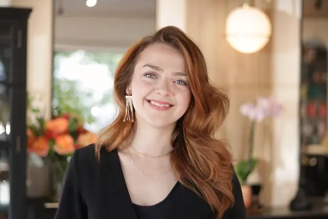 A woman with long auburn hair and drop earrings smiling, orange flowers and orchids behind her.