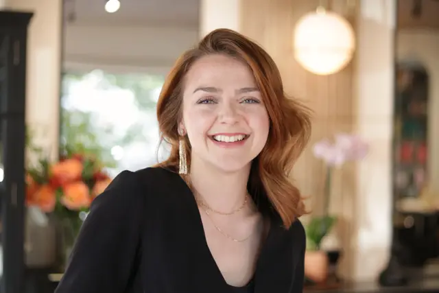 A woman with auburn hair laughing, turned slightly toward the camera, flowers on the counter behind her.
