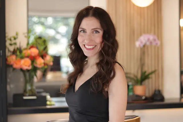 A smiling woman with long dark wavy hair in a black dress, orchids and flowers behind her.