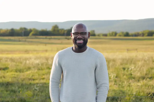 A smiling man in a cream sweater and glasses standing in a sunlit meadow, hills on the horizon.