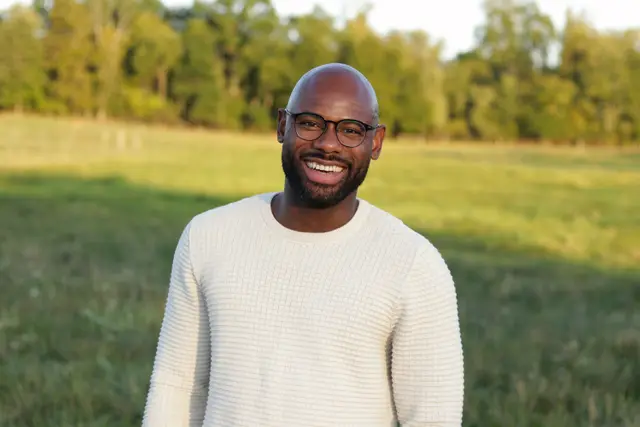 A man in glasses and a cream sweater smiling in a green field at golden hour, trees behind.