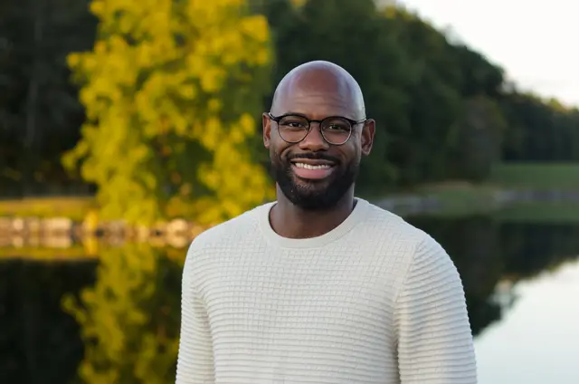 A man in glasses and a cream sweater smiling in front of a pond reflecting yellow-green trees.