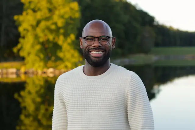 A smiling man in glasses and a cream sweater up close, a pond and trees softly blurred behind.
