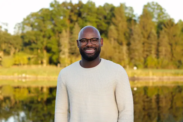 A man in a cream sweater standing by a still pond with trees reflected in it.