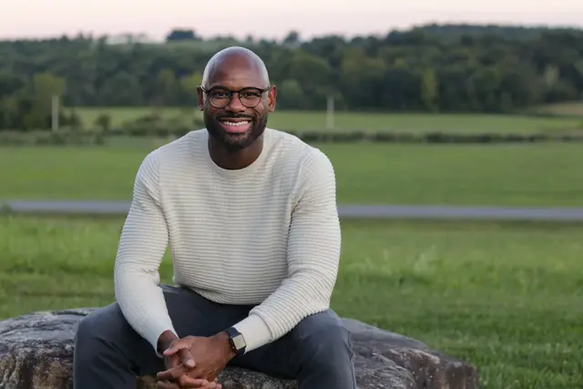 A man in glasses and a cream sweater sitting on a large rock in a field, smiling.