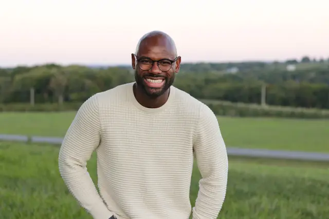 A laughing man in a cream sweater in a field at dusk, a pink sky over the tree line.