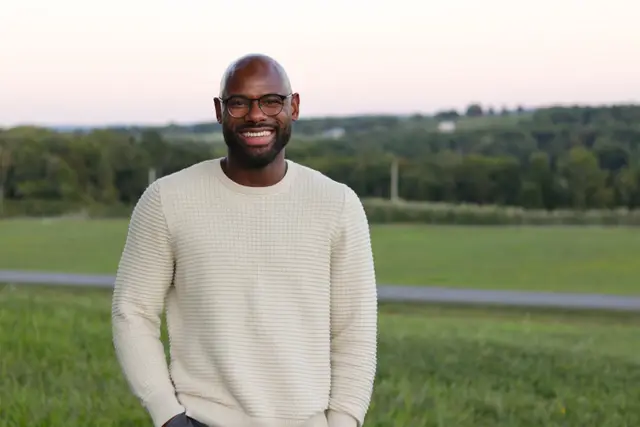 A man in glasses and a cream sweater smiling with his hands in his pockets in a field at dusk.