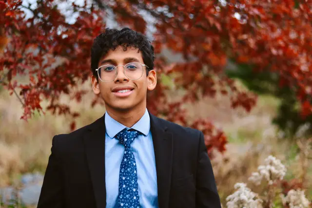 A young man in glasses, a dark suit and a blue tie smiling under a tree with deep red leaves.