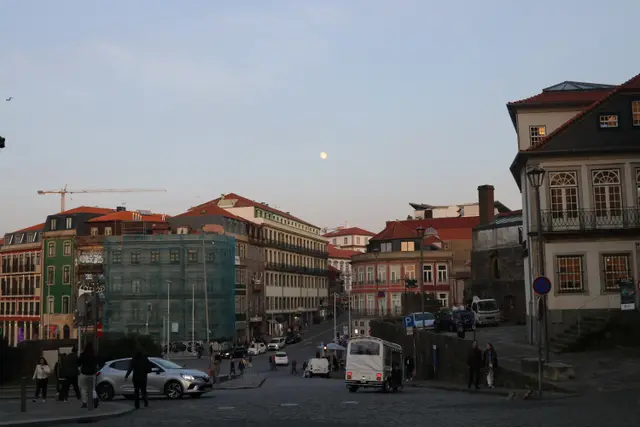 A Porto street at dusk, cars and people below old townhouses and the moon over a construction crane.