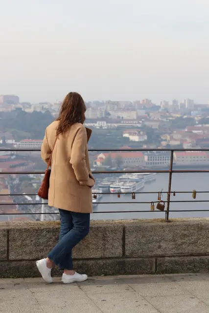A woman in a camel coat leaning on a stone parapet, looking out over the city and river.
