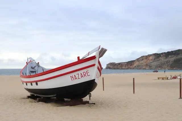 A traditional red and white fishing boat painted Nazaré, propped on the sand, the headland behind.