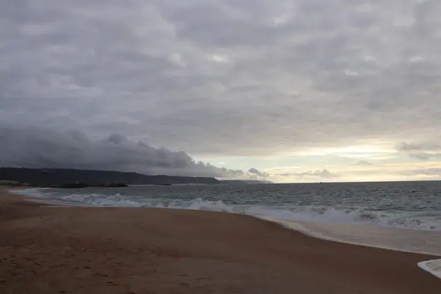 A wide empty beach under heavy cloud, a dark band of rain moving in over the headland.