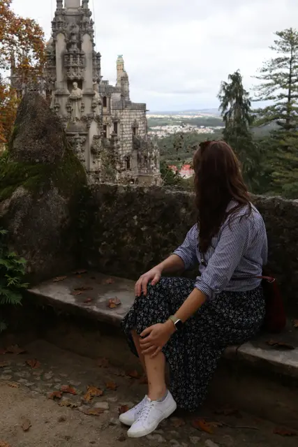 A woman sitting on a stone bench beside the carved gothic tower at Quinta da Regaleira.