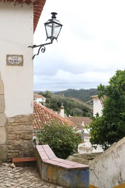 An iron street lamp on a white wall above red roofs and green hills.