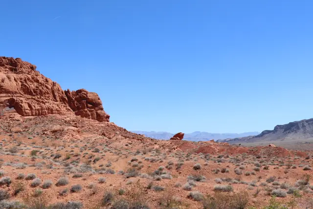 A red rock outcrop above desert scrub under a clear blue sky, mountains on the horizon.