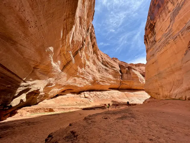 A vast curved sandstone alcove opening to a blue sky, tiny figures on the sand below.