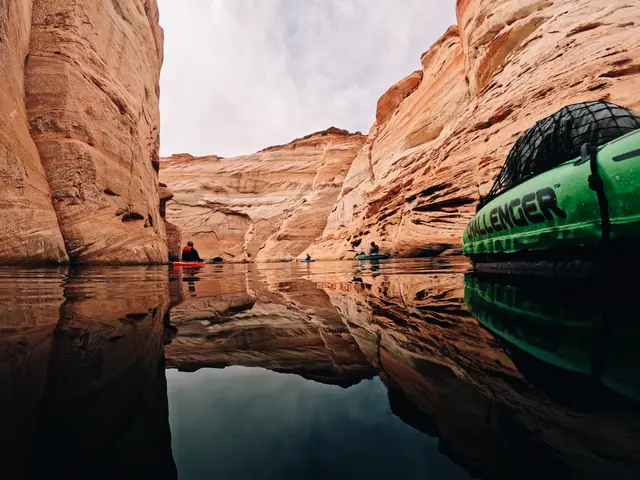 Red sandstone walls reflected in still water, a kayaker in the distance and the bow of a green boat on the right.