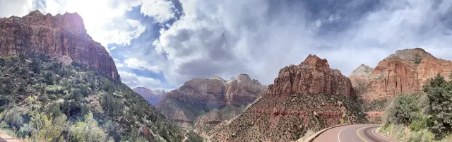 A wide panorama of red cliffs and peaks along a road curving into Zion under a cloudy sky.